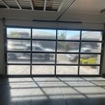 Glass-panel garage door with metal frame, sunlight casting shadows inside, vehicles and houses visible outside.